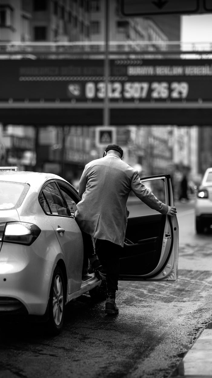 Black and white photo of man in coat exiting a car on a city street.