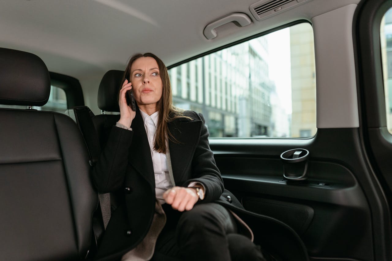 A professional woman in a blazer talks on her smartphone while seated in a car.